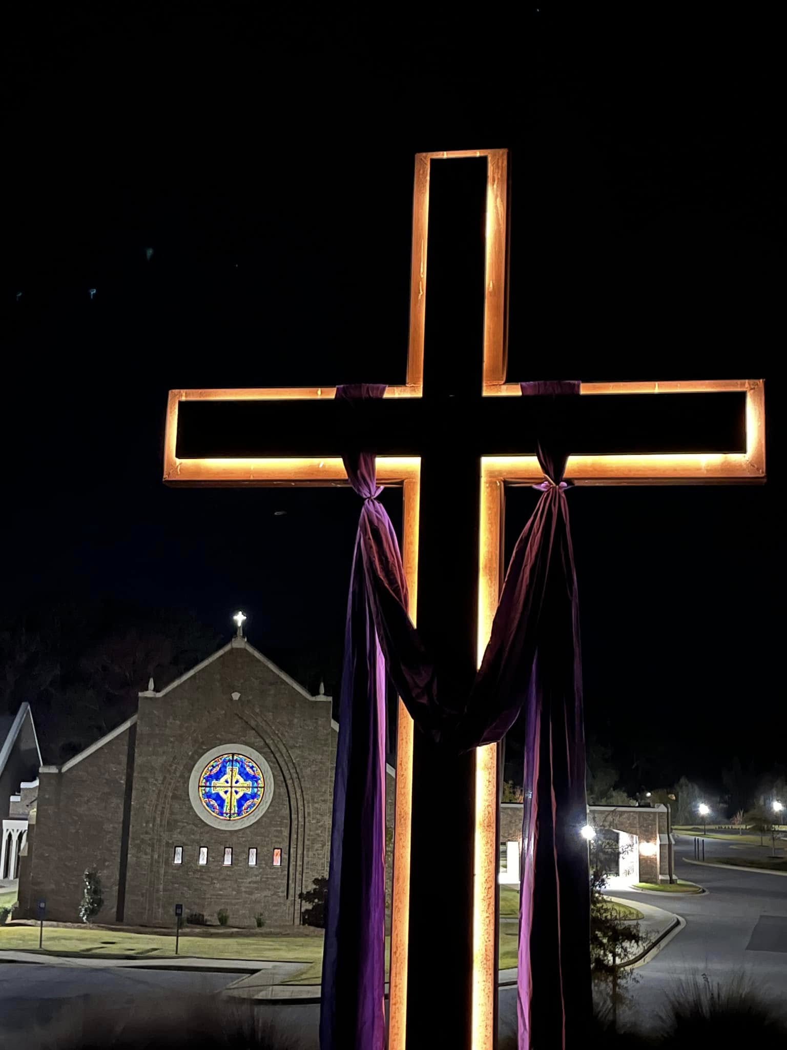 The cross lit at night before the sanctuary, the rose window glowing