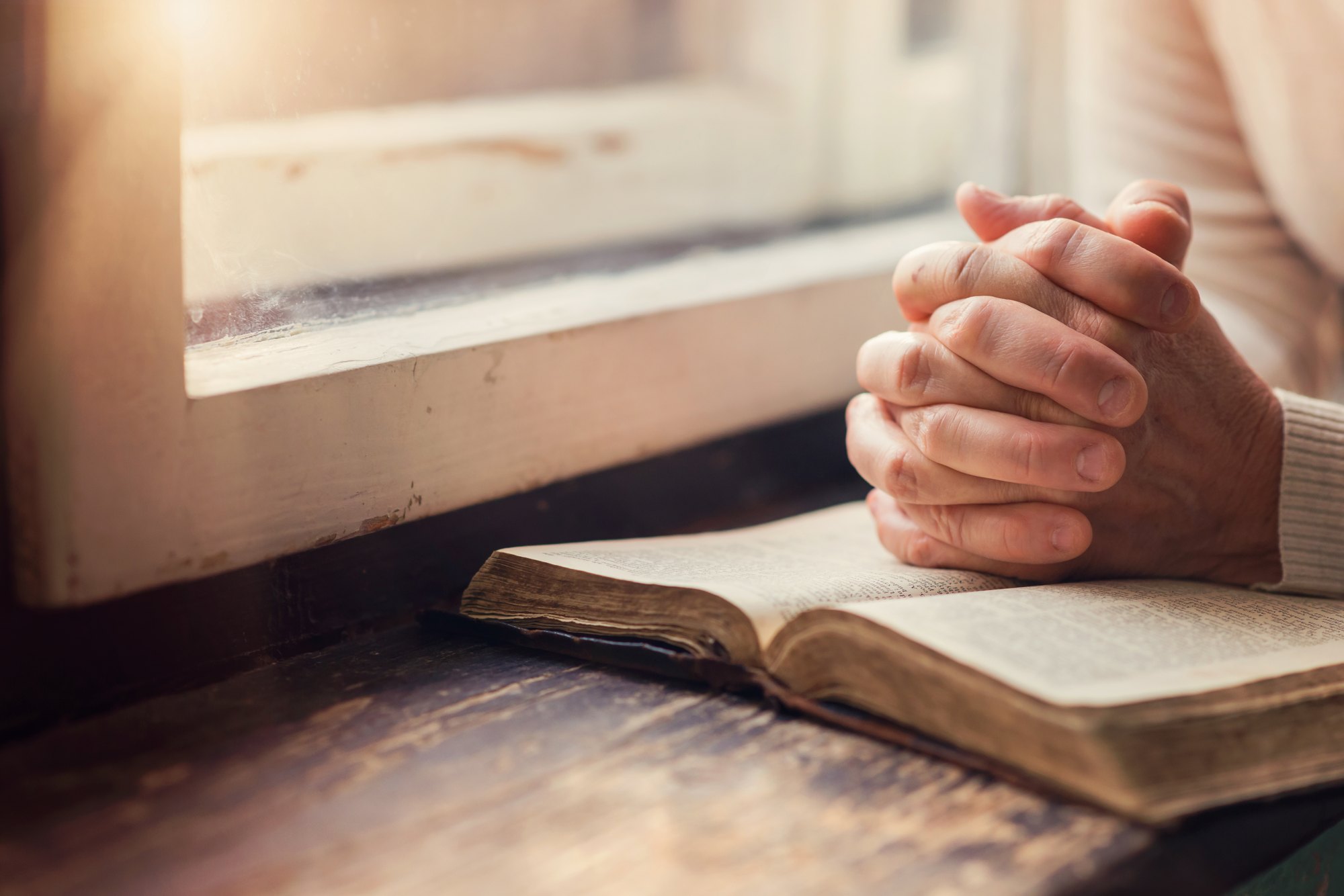 Hands folded in prayer over an open Bible at a window